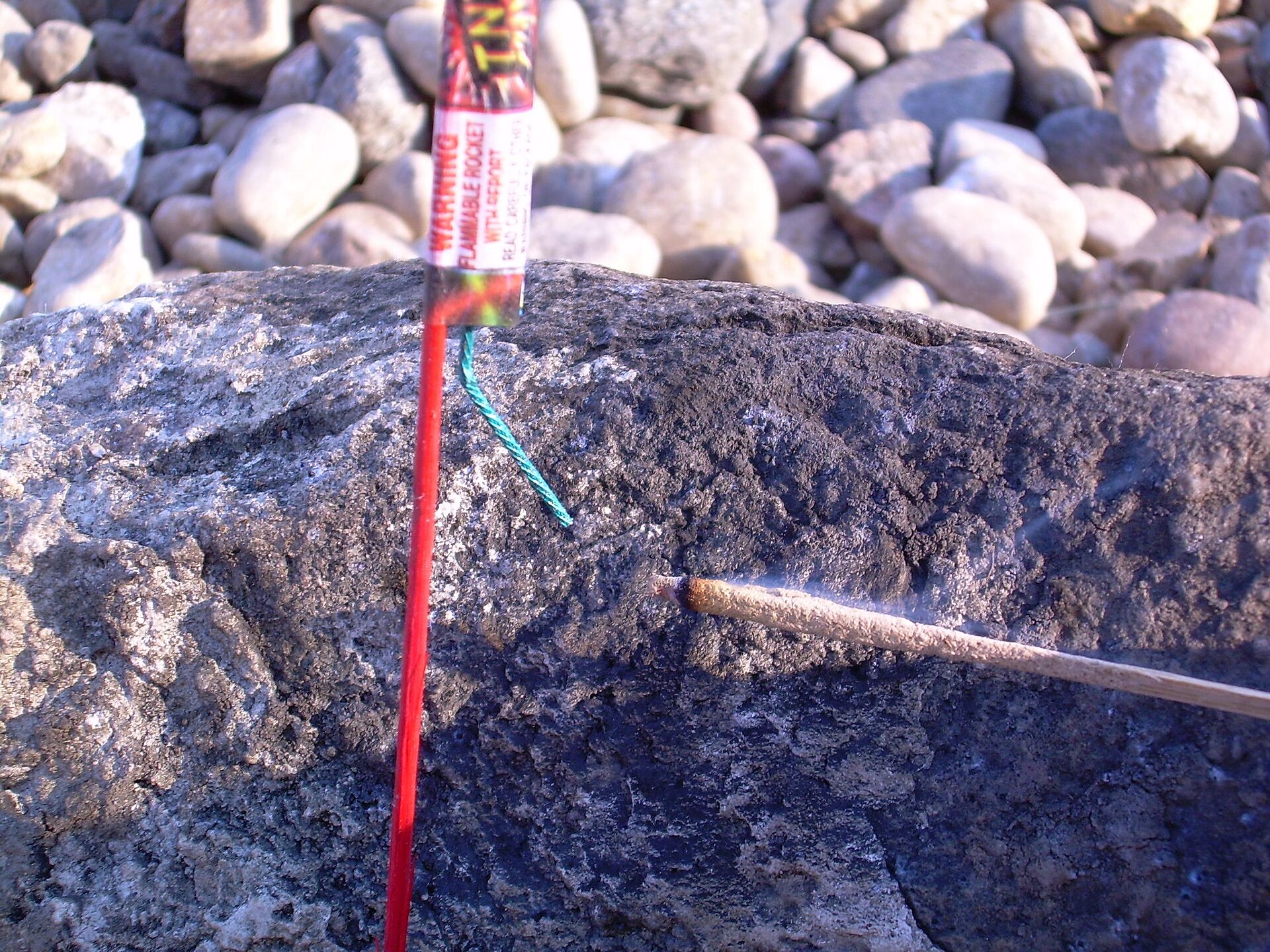 Figure 3 — A stick rocket with a green visco fuse being lit by a smouldering punk on a rock, the fuse trailing loose beside the case.