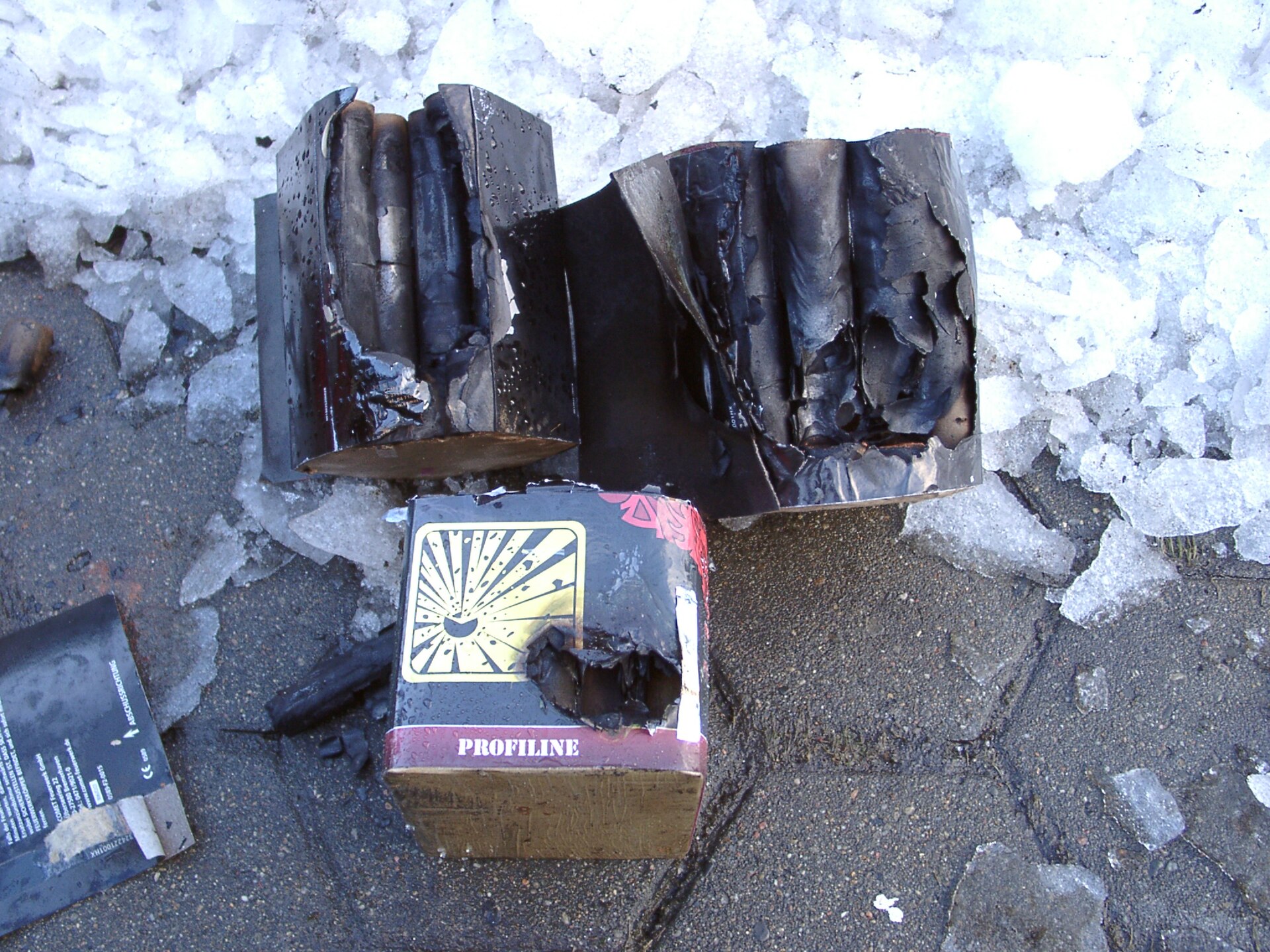 Figure 2 — Burnt-out consumer firework cake tubes lying in slush after use: the outer wrap is charred away in places, showing the bank of thick-walled paper tubes glued side by side, and a separate cardboard …