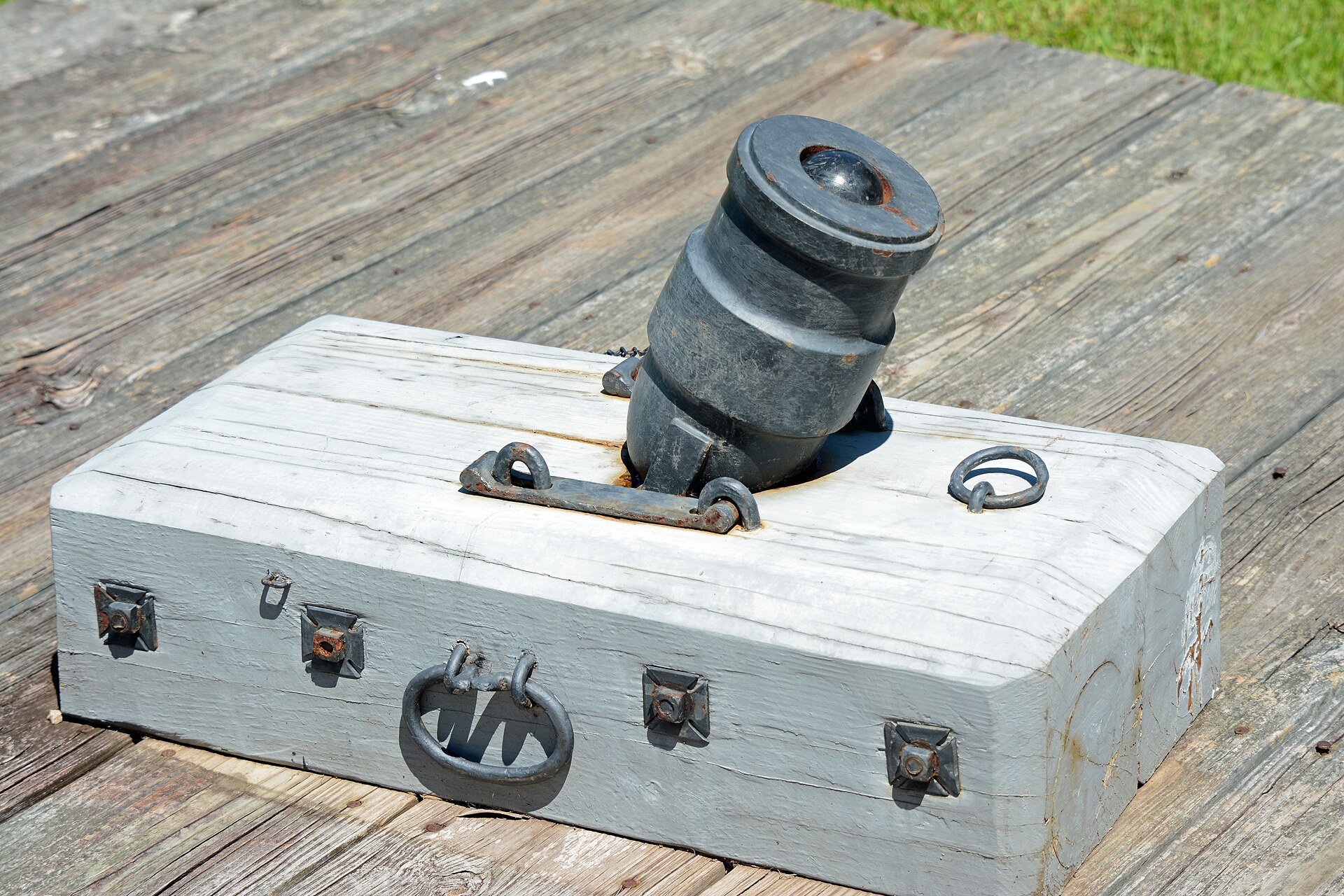 Figure 2 — A coehorn mortar on its heavy timber bed at Fort King George, Georgia: a short, stepped, bell-mouthed bronze tube on trunnions, bolted to a squared block with lifting rings at the corners. This is …