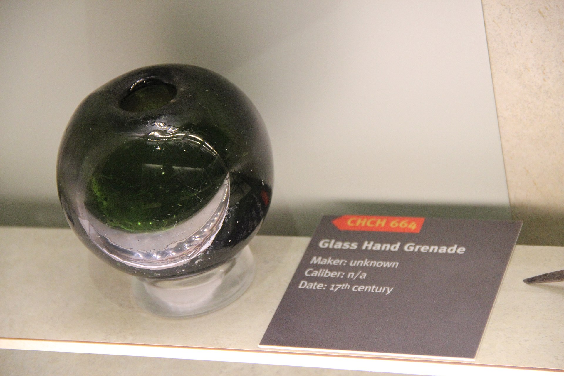 Figure 3 — A seventeenth-century glass hand grenade in a museum case: a heavy dark-green sphere of thick blown glass with a wide opening at the top, catalogued as CHCH 664, maker unknown, date 17th century.