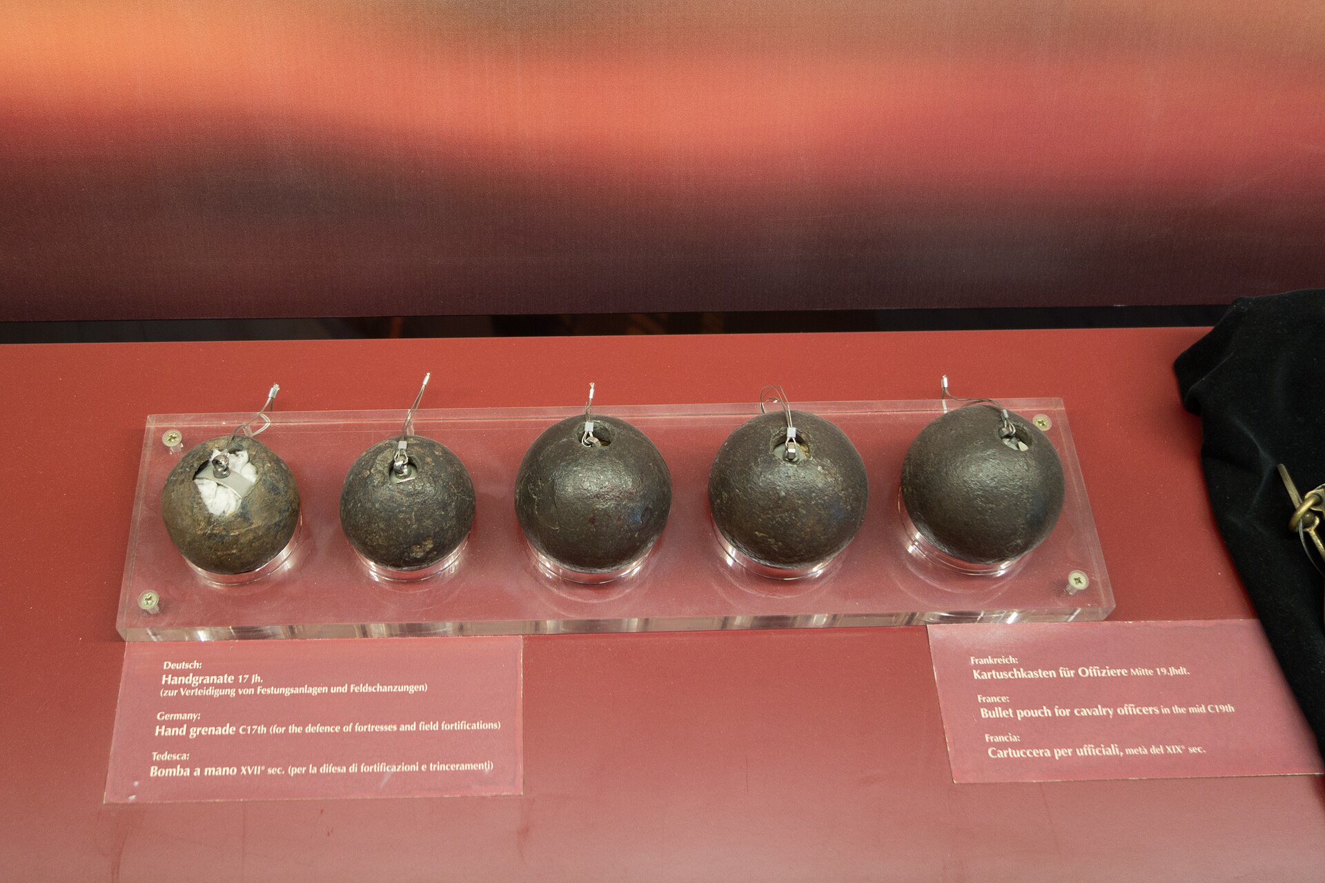 Figure 2 — Five seventeenth-century German iron hand grenades in a museum case, spherical, roughly fist-sized, each with a fuse hole at the top. The leftmost is sectioned to show its filling. The label reads,…