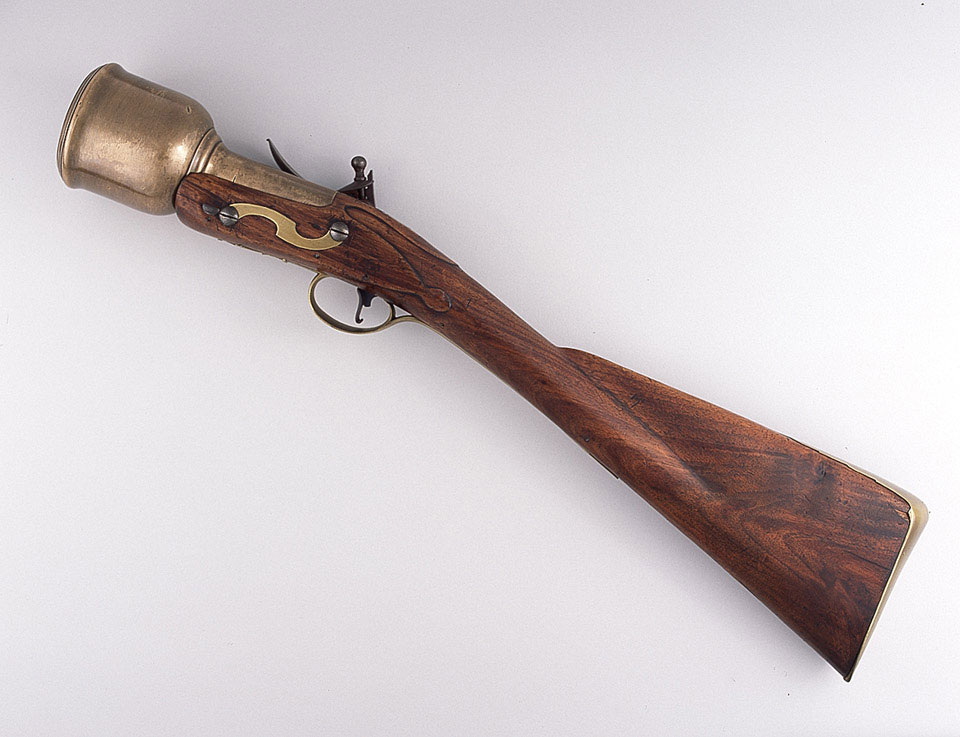 Figure 3 — A British flintlock hand mortar of about 1760 to 1770, photographed from above at an angle against a grey ground: a full walnut stock with a brass butt plate and side plate, a small flintlock, and …