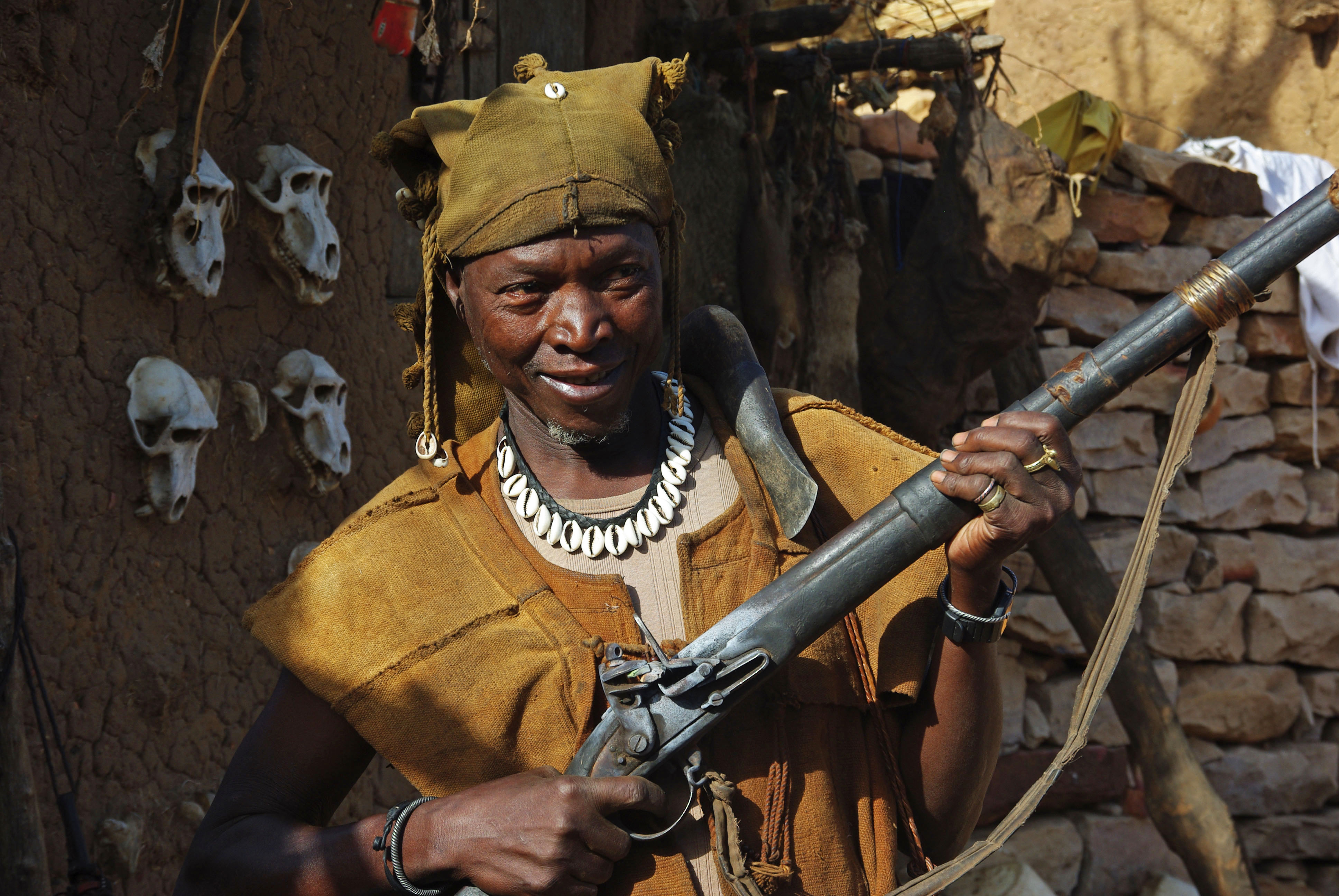 Figure 5 — A present-day Dogon hunter in Mali, in an ochre tunic and cowrie necklace, shoulders a long flintlock "Dane gun," animal skulls on the wall behind him. The trade-gun tradition is not history here —…