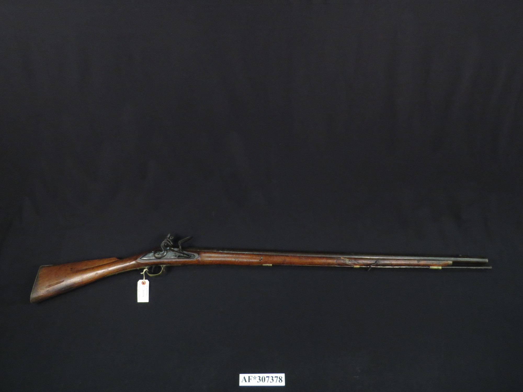 Figure 1 — A full-length British flintlock musket of the Brown Bess pattern, photographed on black: a long smoothbore barrel, a full wooden stock running nearly its whole length, the flintlock at the wrist, a…