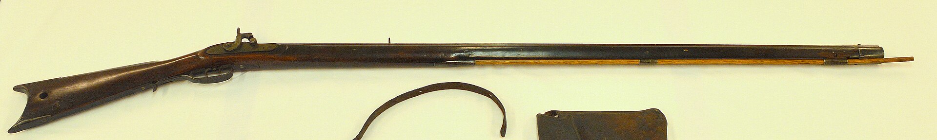 Figure 2 — A half-stock percussion (caplock) American rifle displayed with a powder horn and leather pouch. The lock has no flint or pan — just a hammer falling on a capped nipple at the breech, an all-weathe…