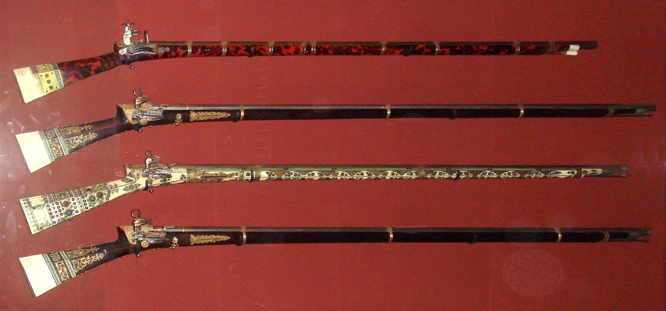 Figure 4 — Four richly decorated Ottoman miquelet tüfek long guns mounted on a red wall — inlaid stocks and long barrels typical of the Mediterranean lock's domain. Source: Wikimedia Commons.