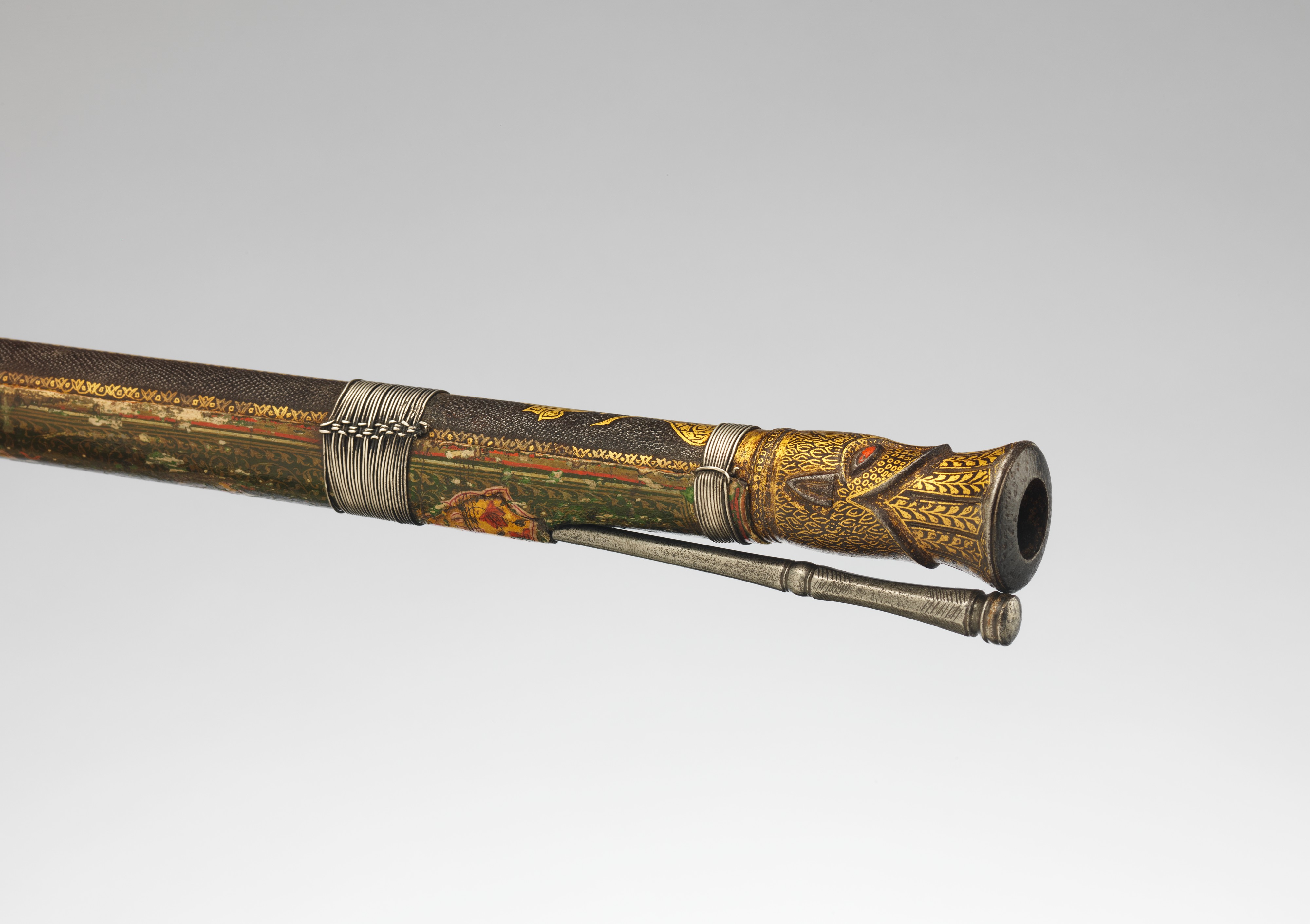 Figure 3 — The muzzle of an Indian toradar matchlock: a gold-decorated barrel ending in a chiseled tiger's-head mouth, with the thin serpentine match-holder alongside. The mechanism is the humble matchlock; t…