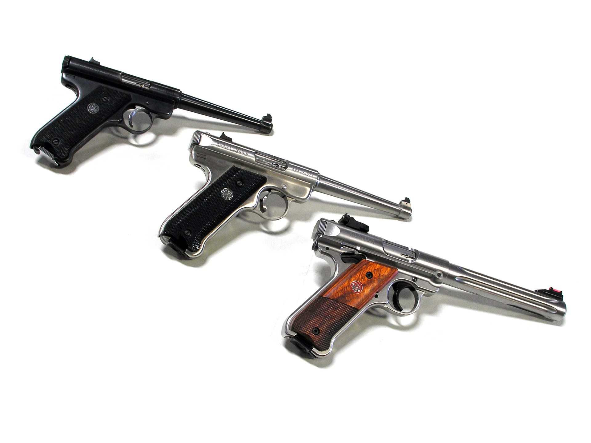 Figure 1 — Three generations in one frame, as photographed and identified by their owner: a Mark I at top, a stainless Mark II in the middle, and a stainless Mark IV Hunter with fluted barrel and laminate gri…