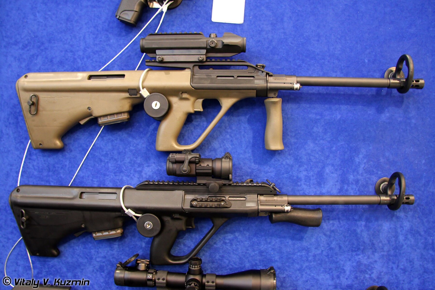 Figure 5 — Two Steyr AUG A3s at the Interpolitex exhibition in Moscow in 2011, in tan and in black, each with a Picatinny flat-top carrying an optic and a rail below the barrel. The serial numbers have been p…