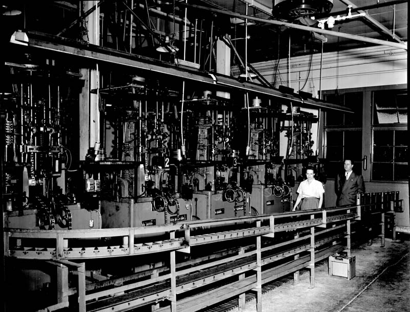 Figure 5 — A bank of numbered automatic machines on the Daisy factory floor, fed from a conveyor that runs the length of the row and driven from an overhead line shaft. Two people are enough to tend four stat…