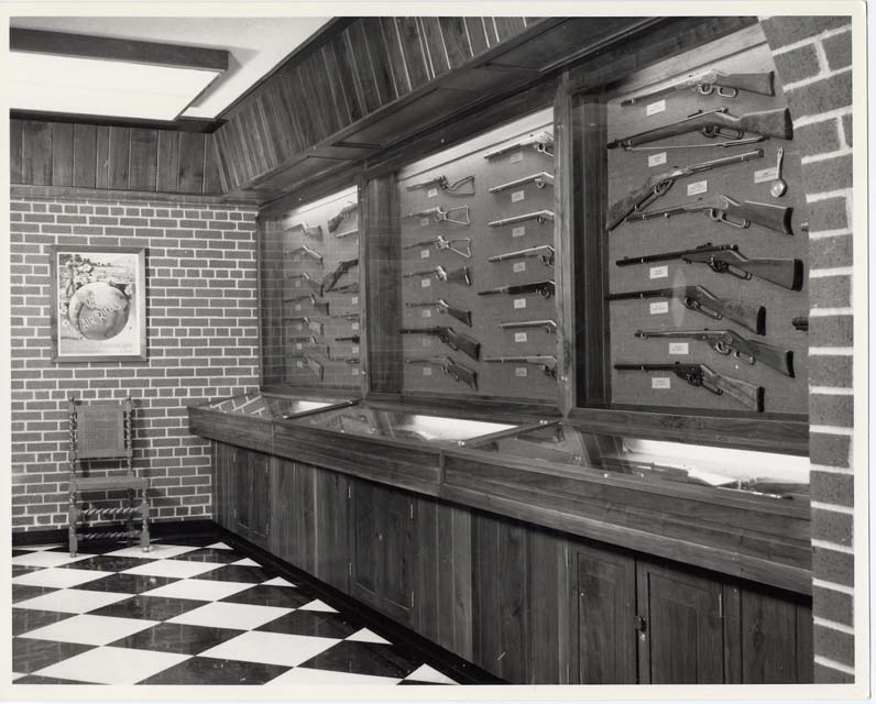 Figure 4 — A Daisy display room, its walls given over to lit cases of air rifles hung in rows and individually labelled, with flat cases beneath. A company that keeps its own back catalogue on the wall like t…